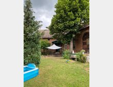 Gartenbereich vor einem Backsteingebäude mit Satteldach, großen Bäumen und einer weißen Sonnensegelüberdachung. Im Vordergrund eine aufblasbare blaue Planschbeckenwanne und eine Rasenfläche.