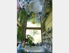 Badezimmer mit einem runden Whirlpool in der Mitte, umgeben von Zimmerpflanzen, Regalen mit Porzellan und einem großen Fenster, das den Blick auf eine grüne Außenlandschaft freigibt. Die Decke ist mit Spiegelfliesen verkleidet, die den Raum optisch vergrößern.