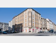 Eckgebäude einer Straße in einem urbanen Gebiet mit grauer Fassade und größtenteils mit Holzplatten verschlossenen Fenstern. Im Erdgeschoss befinden sich Geschäfte mit roten Schildern, und mehrere Autos sind entlang der Straße geparkt.