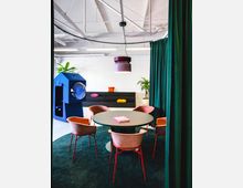 Ein moderner Besprechungsraum mit einem runden Tisch, umgeben von rosafarbenen Stühlen mit roten Beinen, steht auf einem dunkelgrünen Teppich. Im Hintergrund sind ein blaues Möbelstück, eine schwarze Sitzbank mit bunten Kissen, eine Zimmerpflanze und eine weiße Wand zu sehen; von der Decke hängt eine lilafarbene Lampe.