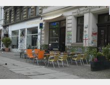 Straßenecke mit einem kleinen Restaurant, dessen Fassade mit grünen Fliesen und Graffiti bedeckt ist, daneben ein Schaufenster. Vor dem Lokal stehen orangefarbene und beige Stühle mit Tischen auf dem Gehweg.
