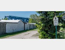 Ein Weg führt an einfachen, weißen Gebäuden entlang, die eine ländliche Umgebung andeuten. Im Hintergrund ist eine blaue Stahlbrücke erkennbar, rechts steht ein Schild mit der Aufschrift „Naturschutzgebiet“ vor einem großen Nadelbaum.