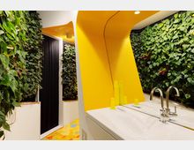 Ein modernes Badezimmer mit einer gelben Wandverkleidung, einem weißen Doppelwaschbecken und glänzenden metallischen Wasserhähnen. Die Wände sind dekoriert mit grünen Pflanzen in vertikalen Begrünungselementen, und ein auffälliger Bodenbelag mit gelbem Muster ergänzt das lebendige Design.