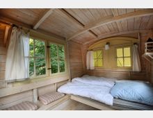 Kleiner Innenraum aus hellem Holz mit gebogener Decke, einem Doppelbett mit weißer Bettwäsche und zwei Fenstern mit Vorhängen, die auf grüne Bäume blicken. Sitzbank mit gestreiften Polstern unter den Fenstern und eine kleine Lampe an der Wand.