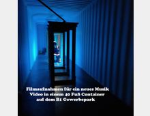 Blick in das Innere eines beleuchteten, blauen Schiffscontainers mit einer Person im Hintergrund, die durch mehrere aneinandergereihte, dunkle Rahmen sichtbar ist. Text auf dem Bild beschreibt Filmaufnahmen für ein Musikvideo im B1 Gewerbepark.