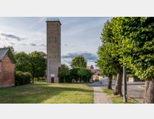 Ein rechteckiger, hoher Turm aus grauem Backstein mit einer Plattform auf der Spitze steht in einer Grünfläche. Links ist eine rote Backsteinmauer mit einem begrünten Dach, rechts eine mit Bäumen gesäumte gepflasterte Straße, und dahinter sind weitere niedrige Gebäude zu sehen.