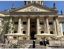 Historisches Gebäude mit klassizistischer Fassade, verziert durch Säulen mit korinthischen Kapitellen und ein Relief im Giebeldreieck. Im Vordergrund stehen Sonnenschirme und Gartenmöbel auf einer gepflasterten Fläche, unter blauem Himmel.