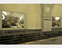 U-Bahn-Station mit stark gewölbter Decke, beige gefliesten Wänden und schwarzen Wandfliesen an der unteren Hälfte. Zwei gerahmte Bilder zeigen lokale Szenen, daneben steht der Schriftzug „Heidelberger Platz“.