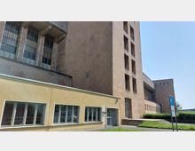 Außenansicht eines großen Gebäudekomplexes mit hellbrauner Steinfassade und großen Fenstern, kombiniert mit einem niedrigeren gelb gestrichenen Anbau mit modernen Glasfenstern. Im Vordergrund ist eine Grünfläche zu sehen, rechts daneben ein Parkplatzschild.