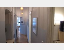 Schmaler, heller Flur in einer Wohnung mit Holzboden, weißen Wänden und blauer Wandunterseite. Der Flur führt zu einem Raum mit einem großen Fenster, während an den Seiten Bilder und Türen sichtbar sind. Im Vordergrund rechts ist ein Teil eines Badezimmers mit Fliesen erkennbar.