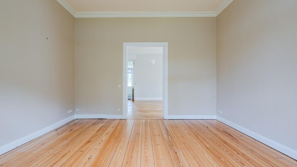 Leerer, heller Wohnraum mit hohen Decken, weißer Stuckleiste und breiten Dielen-Holzboden. In der Mitte führt eine Türöffnung in einen weiteren Raum mit ähnlichem Boden und einem Fenster im Hintergrund.