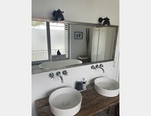 Moderner Badezimmerbereich mit einem großen rechteckigen Spiegel über einer hölzernen Waschtischplatte, auf der zwei weiße, runde Aufsatzwaschbecken mit Wandarmaturen angebracht sind. Im Hintergrund sind ein Fenster mit Sichtschutzfolie, ein gerahmtes Bild und ein Wäschekorb zu sehen.