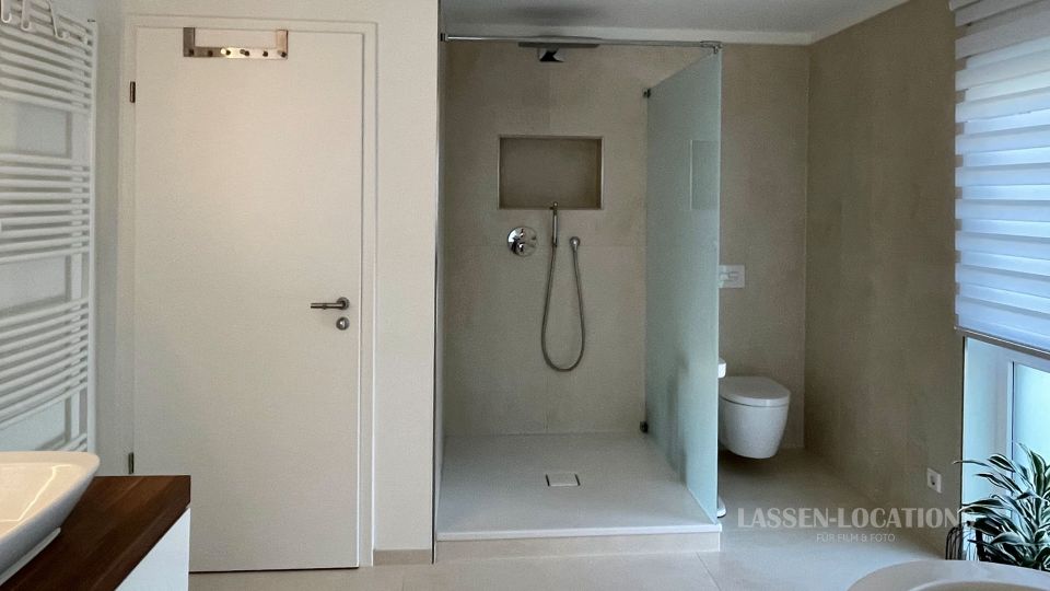 Modernes Badezimmer mit bodengleicher Dusche und Glasabtrennung, wandhängendem WC sowie hellen, neutralen Wand- und Bodenfliesen. Links sind eine weiße Tür und ein Handtuchheizkörper zu sehen, rechts ein großes Fenster mit Rollos und eine Pflanze.