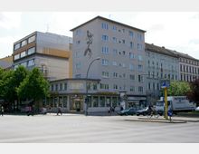 Straßenszene mit einem sechsstöckigen Gebäude im Hintergrund, das mit einem abstrakten Wandmosaik verziert ist. Im Erdgeschoss befindet sich ein Commerzbank-Filiale, daneben eine U-Bahn-Markierung sowie Geschäfte, Autos und Fußgänger an einer Kreuzung.