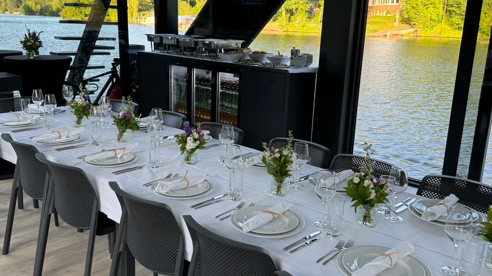 Eleganter Speiseraum mit großen Fenstern, die einen Blick auf einen See und Bäume bieten, ausgestattet mit einem gedeckten Tisch mit weißen Tischdecken, Blumenarrangements und Geschirr; im Hintergrund ein schwarzer Schrank mit Getränkekühlern und Buffetelementen.