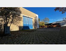 Eine freistehende, beige Gebäudewand mit einer großen blauen Doppeltür, umgeben von einem gepflasterten Hof mit herbstlichem Laub. Im Hintergrund sind kleinere Gebäude, Bäume und ein klarer, blauer Himmel zu sehen.