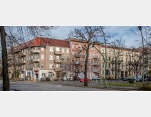 Straßenszene mit mehrstöckigen Wohnhäusern aus der Gründerzeit, inklusive Balkonen und roten Ziegeldächern; die Gebäude stehen an einer belebten Kreuzung mit Fußwegen, Autos und Ampeln. Im Vordergrund sind kahle Bäume und ein Gehweg erkennbar.