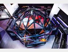 Eine futuristische Innenrauminstallation in Form einer gläsernen, facettierten Kuppel mit schwarzem Metallrahmen, beleuchtet von buntem Licht in Rot, Blau und Lila. Umgeben von modernem Design mit sichtbaren Rohren, weißen Säulen und schräg verlaufenden neonbeleuchteten Wänden.