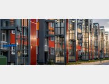 Reihe von mehrstöckigen Wohngebäuden mit bunten Fassaden in Rot und Orange, ausgestattet mit modernen verglasten Außenelevatoren. Vor den Häusern verläuft ein schmaler Gehweg mit Straßenlaternen.