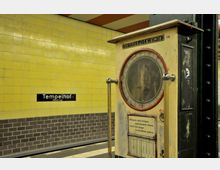 Eine historische Personenwaage mit der Aufschrift „Genaues Gewicht“ steht in einer U-Bahn-Station mit gelben Wandfliesen. Im Hintergrund ist ein schwarzes Stationsschild mit der Aufschrift „Tempelhof (Südring)“ zu sehen.