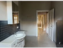 Kleiner Raum mit einem weißen Waschbecken und Unterschrank, gefliester Wand in dunklem Blau und einem Wandspiegel. Eine offene Tür führt in einen angrenzenden Flur mit weiterem Tageslichteinfall und sichtbaren Türen.