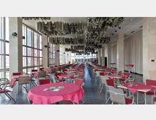 Großer Speisesaal mit vielen Tischgruppen, die mit roten Tischdecken gedeckt sind, und Holzbügenstühlen mit roten Polstern. An der Decke befinden sich dekorative Metallinstallationen, während die Wände große Fenster auf der linken Seite und Vorhänge auf der rechten Seite aufweisen.