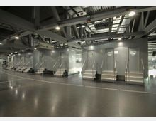 Innenansicht eines modernen Stadions mit einem metallischen Gangbereich. Sichtbar sind mehrere silberne Türen mit runden Fenstern, die über kleine Treppen mit Geländern zugänglich sind, sowie freiliegende Strukturelemente und Beleuchtung an der Decke. Ein Schild markiert den Bereich „Block 18“.