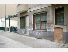 Fassade eines älteren Wohngebäudes mit großen Fenstern und beige-brauner Eingangstür aus Holz; die Wand ist von Graffiti bedeckt. Der Gehweg davor besteht aus Kopfsteinpflaster, und links ist ein Zaun mit Blick auf einen Innenhof zu sehen.