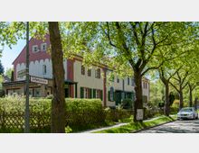 Reihenhäuser in einer Wohnstraße mit hell verputzten Fassaden, grünen Fensterläden und roten Zierelementen, umgeben von niedrigen Hecken und Bäumen, die dichten Schatten auf die gepflasterte Straße werfen. Im Vordergrund steht ein Straßenschild, und rechts ist ein geparktes Auto sichtbar.