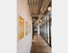 Ein moderner Büroflur mit Holzboden, weißen Wänden und verglasten Bürofronten auf der rechten Seite. Die Decke zeigt sichtbare Rohrleitungen und Kabelkanäle, und an der linken Wand hängen gelb gerahmte Bilder.