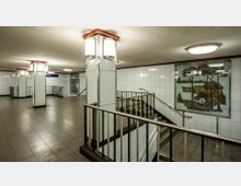 Großzügiger U-Bahn-Zugang mit weißen, gefliesten Wänden, metallenen Geländern und breiten Treppen. Ein großes Wandbild mit einer Landschaftsszene schmückt den Bereich.