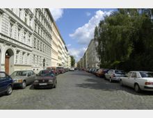 Kopfsteinpflasterstraße in einer Wohngegend mit historischen, mehrstöckigen Gebäuden auf beiden Seiten. Fahrzeuge sind entlang der Straße geparkt, und rechts befindet sich dichter, grüner Baumbewuchs.