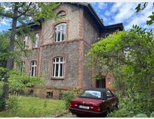 Mehrstöckiges Wohnhaus mit rau verputzter Fassade und roten Ziegelrahmen um die Fenster, teils mit Efeu bewachsen. Davor ein verwilderter Garten mit Bäumen und Sträuchern sowie ein geparktes rotes Cabrio nahe dem Hauseingang.
