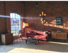 Ein gemütlicher Raum mit rohen Backsteinwänden und großen Fenstern, durch die Tageslicht fällt. Der Raum ist mit einem roten Sofa, einem Sessel, einer Stehlampe, Holzmöbeln und einem kleinen Couchtisch ausgestattet. Wanddekoration und ein Schild ergänzen das rustikale Ambiente.