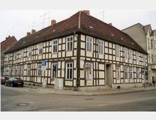 Fachwerkhaus an einer Straßenecke mit weißer Putzfassade und dunklen Holzbalken. Das Gebäude hat ein rotes Ziegeldach, zahlreiche Fenster und liegt in einer urbanen Umgebung mit geparkten Autos und Verkehrsschildern.