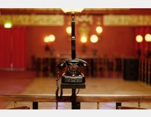 Ein schwarzes Wählscheibentelefon ist auf einem Metallgestell befestigt, im Hintergrund ein großer Raum mit roten Wänden, goldenen Verzierungen, roten Vorhängen und warmem Licht von mehreren kugelförmigen Lampen.