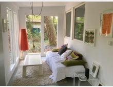 Helles Zimmer mit großen Glasfenstern, die den Blick in einen bewaldeten Garten ermöglichen. Ein weißes Sofa mit bunten Kissen steht neben einem niedrigen Tisch auf einem cremefarbenen Teppich, flankiert von einer roten Stehlampe und minimalistischen Wandbildern.