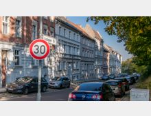 Straßenszene in einer Wohngegend mit mehrstöckigen Altbauten in verschiedenen Farben, parkenden Autos am Straßenrand und einem Verkehrsschild für Tempo 30. Rechts befinden sich Bäume, die einen grünen Bereich andeuten.