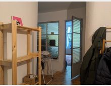 Ein schmaler Wohnungsflur mit Holzboden, einem offenen Holzregal links, und einer Glastür mit strukturierter Verglasung, die zu einem weiteren Raum mit Fenster und Heizkörper führt. Rechts hängen Mäntel auf einer Garderobe.