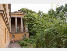 Gebäude im klassizistischen Stil mit einer Vorhalle, die von vier säulengetragenen Portiken geprägt ist, umgeben von dichter Vegetation. Die Fassade aus gelblichem Stein zeigt Fenster und architektonische Details, während der Bewuchs den Eindruck eines überwucherten Gartens vermittelt.