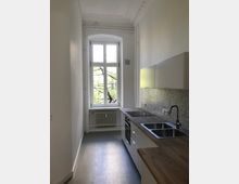 Schmale, moderne Küche mit weißen Wänden und glattem grauen Boden. Auf der rechten Seite befinden sich eine Arbeitsplatte aus Holz, ein Waschbecken, ein Herd und Hängeschränke; links ein großes Fenster mit Blick auf grüne Zweige und darunter ein weißer Heizkörper.