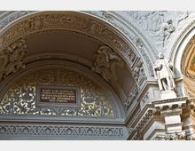 Detailaufnahme eines reich verzierten Kircheninnenraums mit einem kunstvoll gestalteten Rundbogen. Goldene Ornamentik, ein eingearbeitetes Bibelzitat in deutscher Sprache und eine weiße Statue auf einem Podest zieren die Architektur.