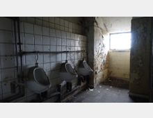 Ein verlassener und verfallener Toilettenraum mit drei Urinalen an einer gefliesten Wand. Die Wände sind teilweise beschädigt und mit abblätternder Farbe bedeckt, ein kleines Fenster lässt schwaches Licht herein.