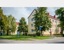 Mehrfamilienhäuser in einem Wohngebiet mit gelber Fassade und roten Ziegeldächern, umgeben von Bäumen und Rasenflächen. Im Vordergrund befinden sich ein Gehweg, ein Straßenschild sowie ein kleines Hinweisschild auf einer grünen Wiese.