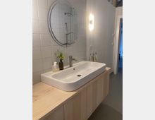 Modernes Badezimmer mit einem breiten, rechteckigen Waschbecken auf einem hölzernen Unterschrank. Darüber hängt ein runder Spiegel an einer gefliesten Wand, daneben eine beleuchtete Wandleuchte.