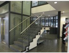 Innenraum eines modernen Gebäudes mit einer Treppe aus dunklem Stein und Geländern aus Metall. Links befindet sich ein Aufzug mit einem Symbol für Toiletten, rechts ein Verkaufsautomat; Glaswände und große Fenster im Hintergrund lassen Tageslicht herein.