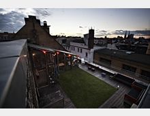 Dachterrasse mit einer kleinen Rasenfläche, umliegendem Holzdeck und Sitzgelegenheiten, teilweise überdacht von einer verglasten Konstruktion. Im Hintergrund sind die Dächer der umliegenden Gebäude und eine Abendhimmel-Szenerie zu sehen.