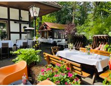 Außenterrasse eines Restaurants mit Holzbänken und weiß gedeckten Tischen, umgeben von blühenden Pflanzen und grüner Vegetation. Im Hintergrund sind ein Fachwerkgebäude, ein Pavillon und Strandkörbe sichtbar, die einen gemütlichen Gartenbereich schaffen.