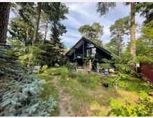 Ein schwarzes Haus mit Satteldach und großer Glasfront, umgeben von einem dicht bewachsenen Garten mit Rasen, Bäumen und vereinzelten Gartenmöbeln. Der Standort wirkt von einem Wald oder einer natürlichen Umgebung umgeben.