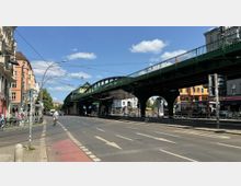 Straßenszene in einer Stadt mit einer erhöhten grünen Brücke, die als Bahntrasse dient. Im Vordergrund sind Fahrradwege, Gehwege und eine mehrspurige Straße, flankiert von Altbauhäusern und Geschäften, sowie vereinzelten Fußgängern und Radfahrern.